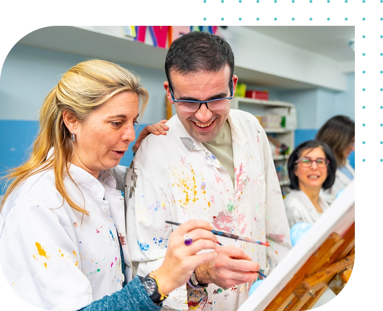Two people wearing paint-splattered lab coats enjoy painting in a classroom, supported by Disabilities Support Services, while a third person sits and observes in the background.